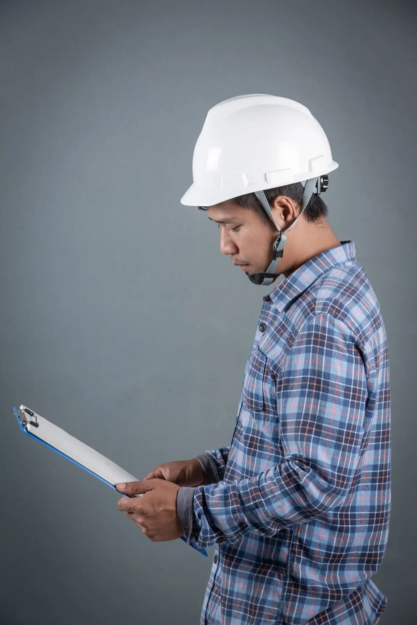 Инженер держит чертежи и читает заметки на планшете с зажимом, стоя на сером фоне.