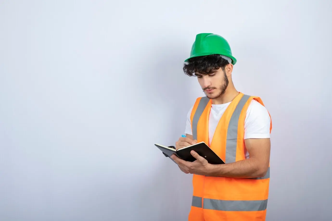 Привлекательный мужчина-инженер в каске пишет в блокноте на фоне белой стены. Фотография высокого качества.